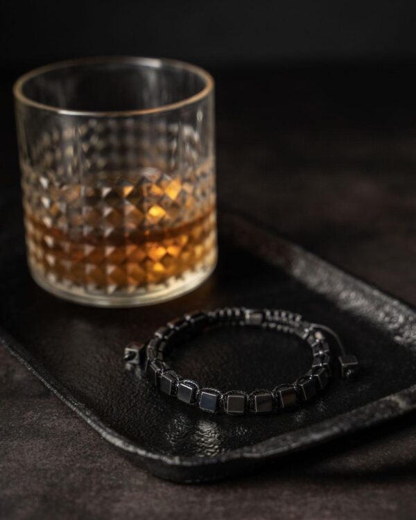 Szklanka bursztynowego napoju obok czarnej bransoletki na ciemnej tacy.