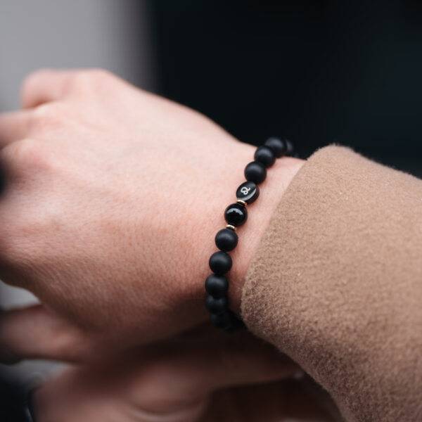 Czarna bransoletka z okrągłych koralików noszona na nadgarstku osoby ubranej w beżowy płaszcz.