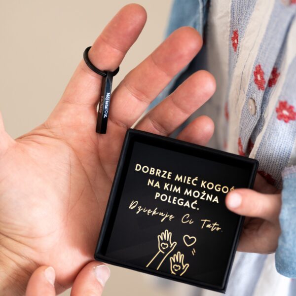 Osoba trzyma brelok z napisem 'Mój Tata' i pudełko z tekstem 'Dobrze mieć kogoś, na kim można polegać. Dziękuję Ci Tato.'