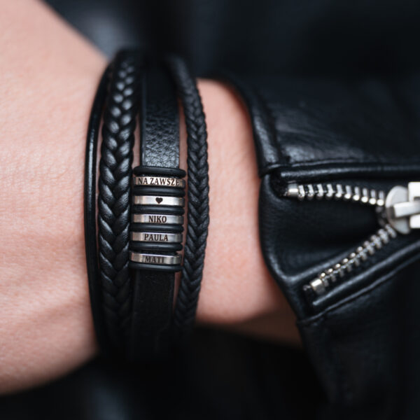 Czarna skórzana bransoletka z wygrawerowanymi imionami: Niko, Paula, Matti oraz napisem 'Na zawsze', trzymana na ręce w skórzanej kurtce.