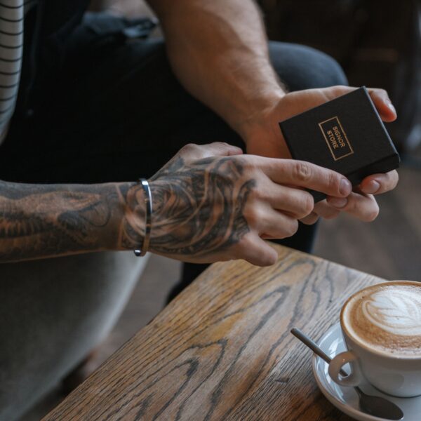 Osoba z tatuażem na przedramieniu trzyma czarne, małe pudełko obok filiżanki kawy na drewnianym stole.