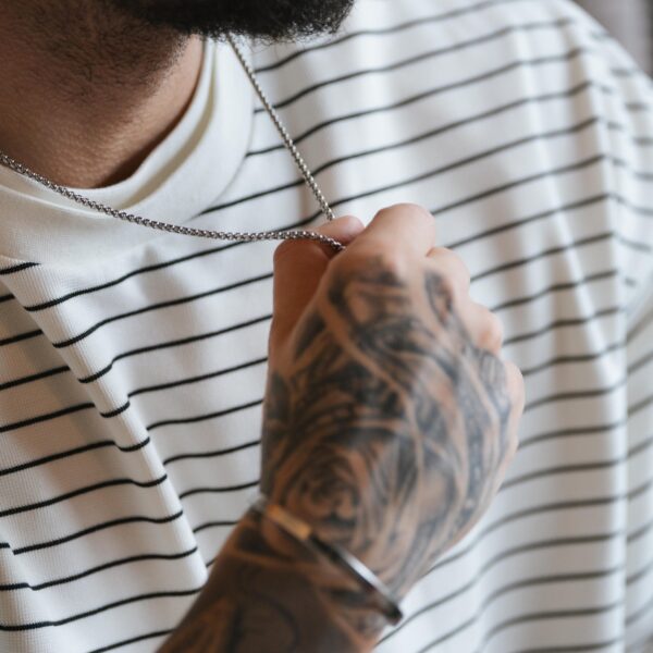 Osoba w białej koszulce w paski trzymająca srebrny łańcuszek, z tatuażami na dłoni.