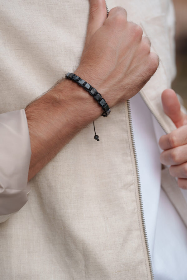 Zbliżenie na męski nadgarstek z czarną bransoletką, osoba reguluje beżową kurtkę.