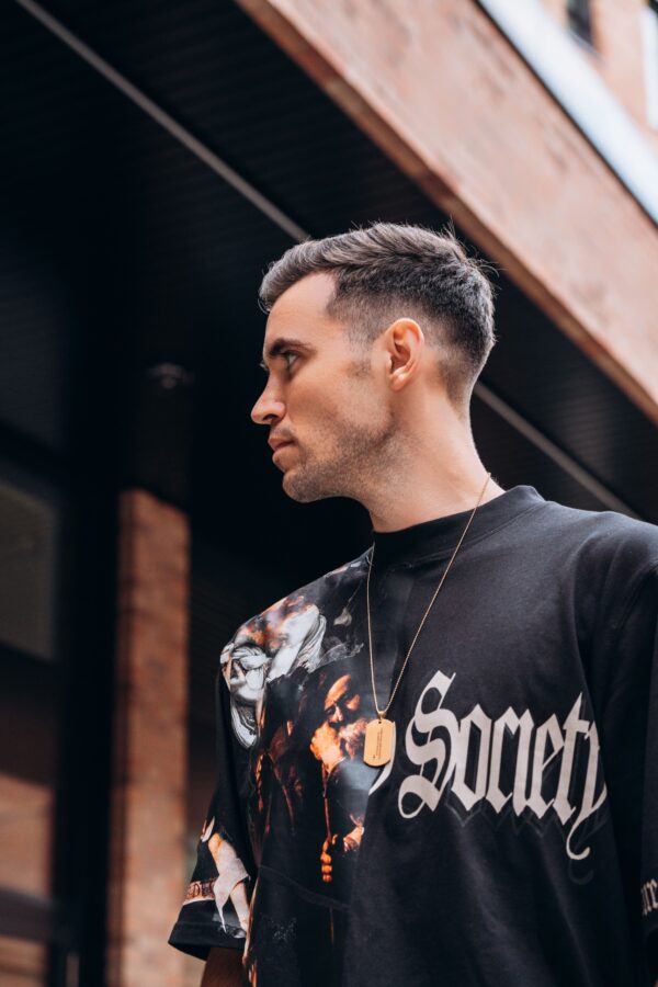Mężczyzna w czarnej koszulce z napisem 'Society' patrzy w bok na tle ceglanej ściany.