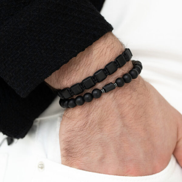Męska ręka z dwoma czarnymi bransoletkami na tle białego ubrania.
