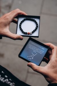 Osoba trzyma pudełko z bransoletką z czarnych koralików i jednym czerwonym koralikiem.