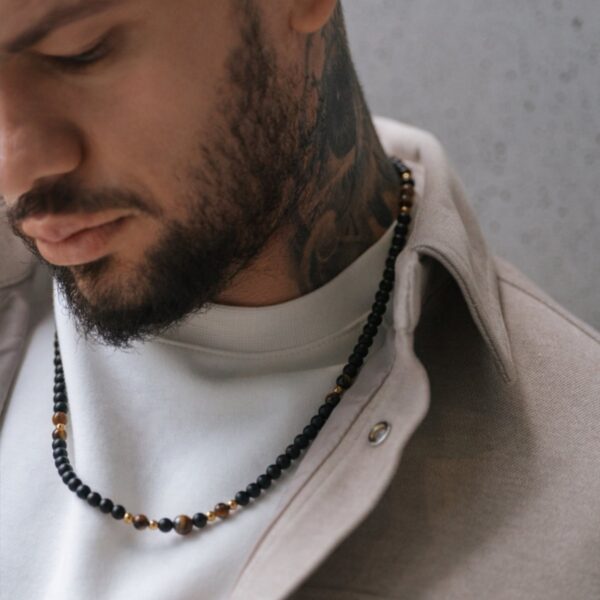 Mężczyzna z brodą, noszący naszyjnik z czarnych koralików i beżową koszulę.