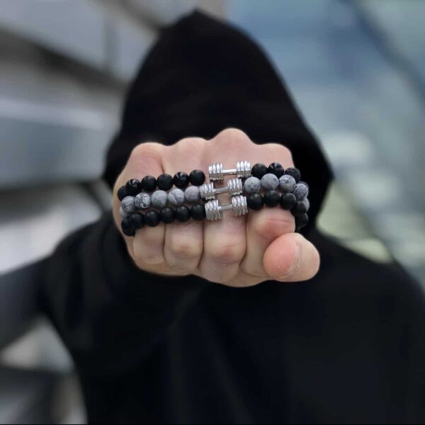 Osoba w ciemnym ubraniu pokazuje pięść z bransoletkami z koralików i hantelkiem.