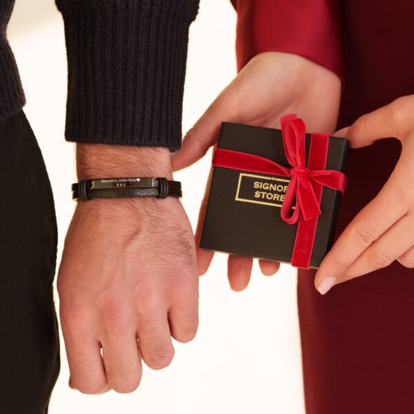 Osoba zakładająca czarną bransoletkę z grawerem i trzymająca zapakowane pudełko z czerwoną wstążką.
