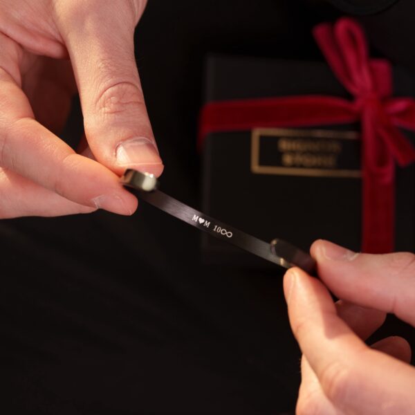 Osoba trzymająca opaskę z napisem 'MOM ♥ 100∞' na tle czarnego pudełka z czerwoną wstążką.