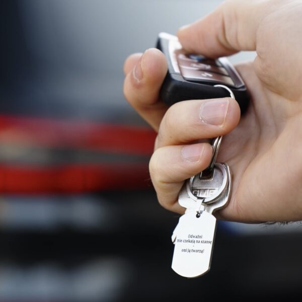 Dłoń trzymająca kluczyk samochodowy z breloczkiem z napisem: 'Odważni nie czekają na szansę, oni ją tworzą.'