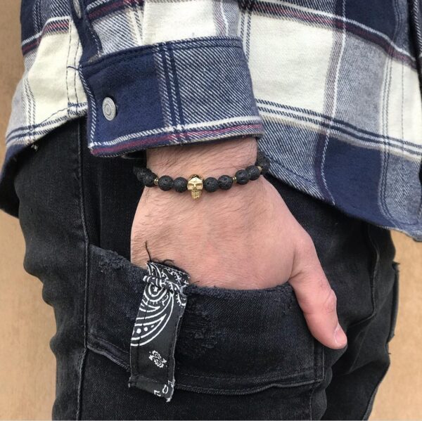 Mężczyzna w koszuli w kratę nosi bransoletkę z czaszką i trzyma rękę w kieszeni jeansów.