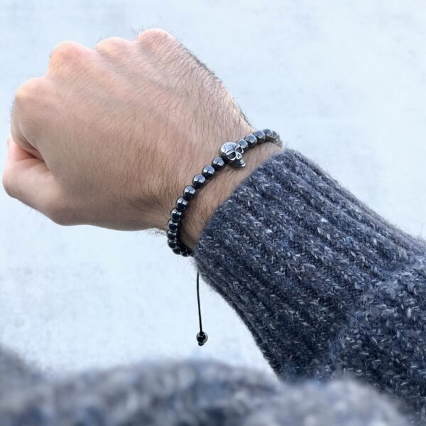 Ręka z ozdobną bransoletką z koralików i ozdobą czaszki, ubrana w szary sweter.