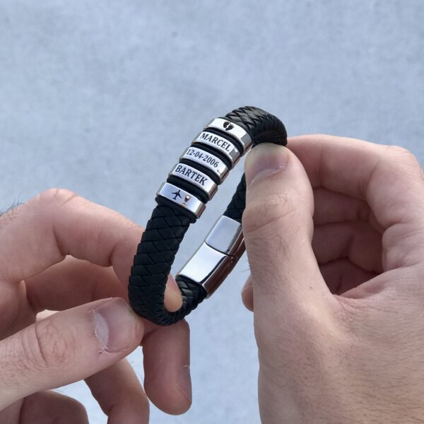 Osoba trzyma skórzaną bransoletkę z metalowymi zawieszkami z wygrawerowanymi imionami i datą.