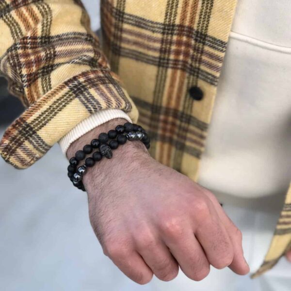 Ręka z czarnymi koralikowymi bransoletkami na tle żółto-brązowej koszuli.