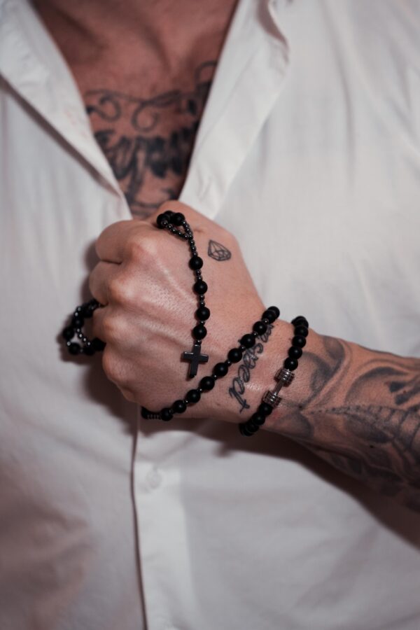 Mężczyzna w białej koszuli z tatuażami na dłoni i czarnym różańcem owiniętym wokół ręki.