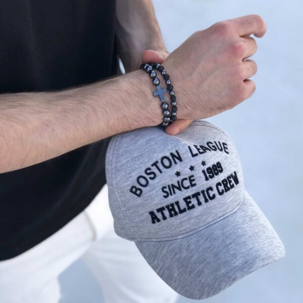 Mężczyzna trzymający czapkę z napisem 'Boston League' i bransoletką z krzyżem.
