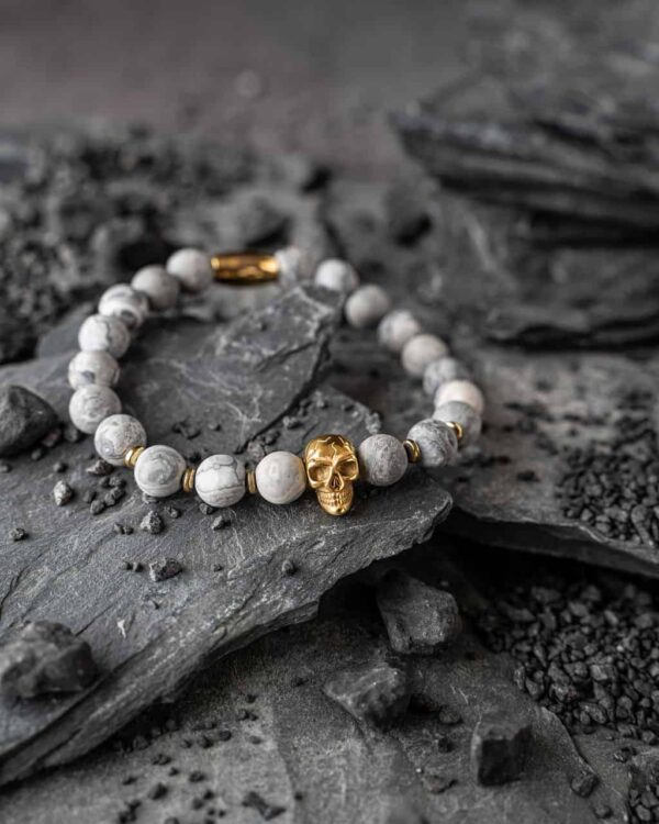 Bransoletka z jasnymi koralikami i złotą czaszką na tle ciemnych kamieni.