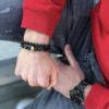 Człowiek w czerwonej bluzie pokazujący rękę z czarnymi koralikowymi bransoletkami i czarnym zegarkiem.