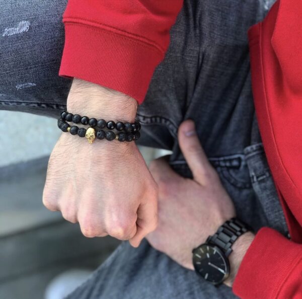 Człowiek w czerwonej bluzie pokazujący rękę z czarnymi koralikowymi bransoletkami i czarnym zegarkiem.