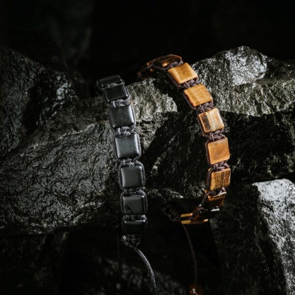 Dwie bransoletki z kwadratowymi koralikami na tle ciemnych skał.