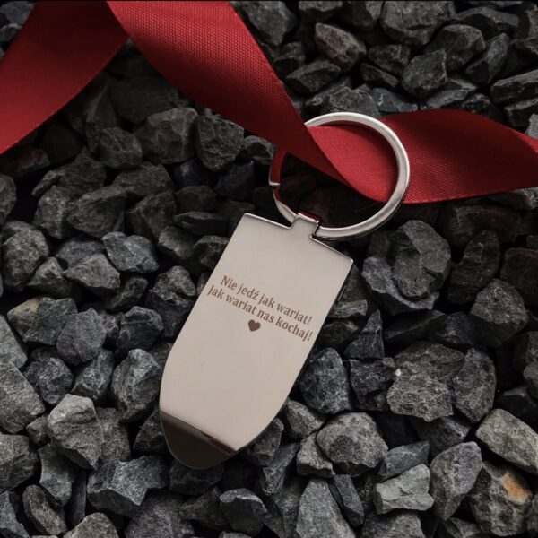 Brelok do kluczy z grawerem 'Nie jedź jak wariat! Jak wariat nas kochaj!' na tle kamieni.