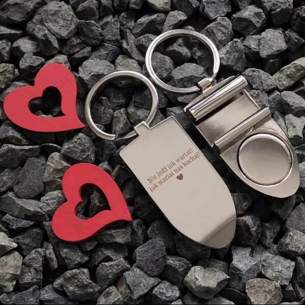 Dwa breloki na żwirze, z czerwonymi sercami obok. Na jednym z nich wygrawerowany napis: 'Nie jeźdź jak wariat, jak wariat nas kochaj!'