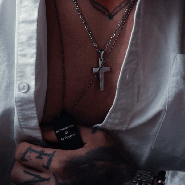Zbliżenie na otwartą koszulę z widocznym naszyjnikiem krzyżem i czarną zawieszką z napisem 'Be Phenomenal Or Be Forgotten'.