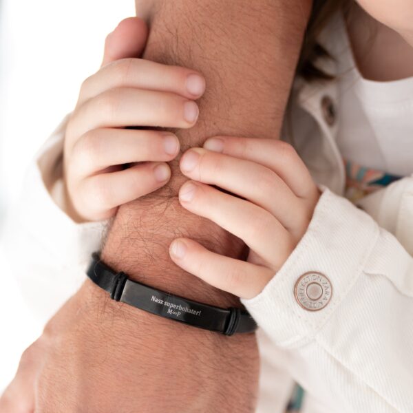 Dziecko trzymające rękę dorosłego z czarną bransoletką z napisem 'Nasz superbohater!'