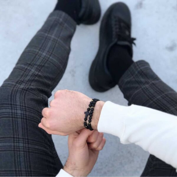 Osoba siedząca w kraciastych spodniach i czarnych butach, nosząca czarną bransoletkę na nadgarstku.