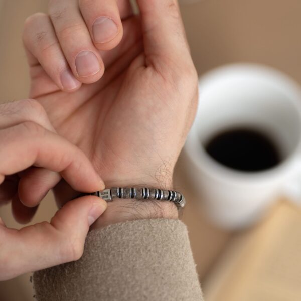 Osoba zakładająca srebrną bransoletkę z czarnymi pierścieniami na nadgarstek, w tle biała filiżanka z kawą.