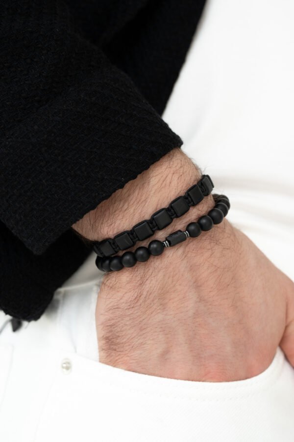 Zbliżenie na męską dłoń z czarnymi bransoletkami na tle białych spodni.