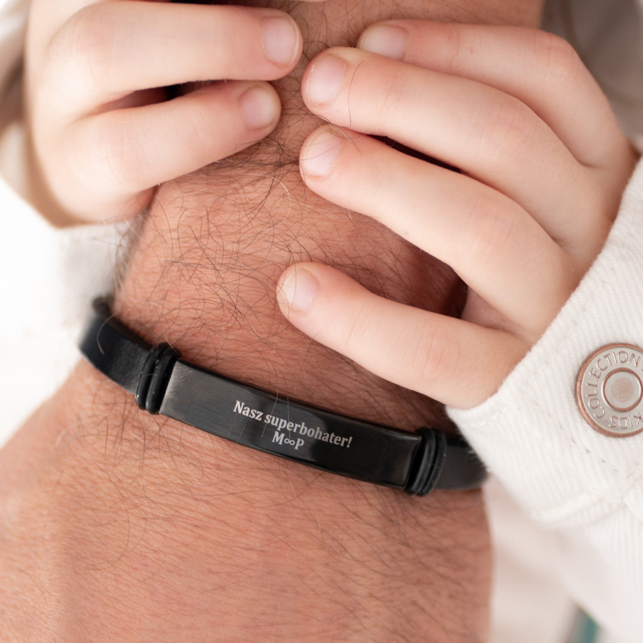 Dłoń dziecka na nadgarstku dorosłego z bransoletką z napisem 'Nasz superbohater!'.