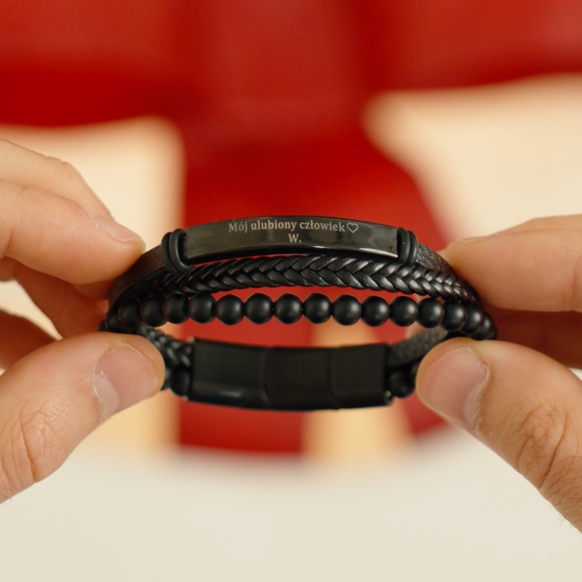 Potrójna bransoletka męska z czarną blaszką, trzymana w dłoniach na czerwonym tle.